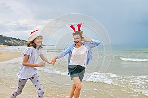 Happy mom and daughter child in Santa hat running along the beach