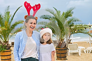 Happy mom and daughter child in Santa hat walking along beach