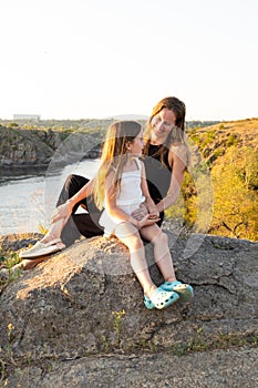 Happy mom and daughter relaxing on rocks in nature against the backdrop of the river. Happy Motherhood, Mother& x27;s Day