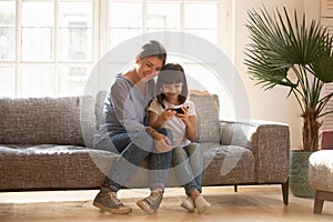 Happy mom and kid daughter using smartphone sitting on couch