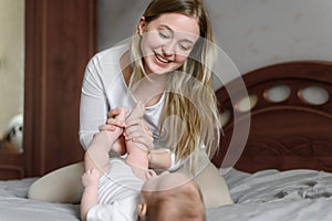 Happy mom plays with her child lying on bed, waking up in morning, smiling, kissing, holding baby legs, enjoying