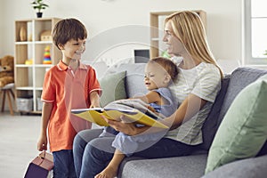 Happy mom reading book of fairy tales to her little children sitting on comfortable sofa at home