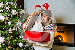 Happy mom with reindeers horns on the top of the head opening the gift with ragdoll cat by the Christmas tree and shining fireplac