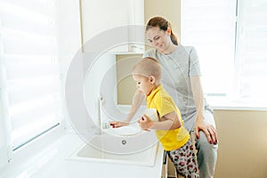 Happy mom teaches little son to wash dishes in the sink.