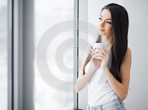 Happy morning. Portrait of beautiful young woman standing at window, drinking coffee and looking outside enjoys of rest