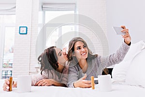 Happy morning of two joyful attractive girls making selfie on white bed. Pretty young women having fun together, smiling