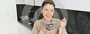 Happy mornings. Gorgeous young woman eating cereals with milk, standing in kitchen with breakfast bowl, enjoying start