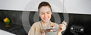 Happy mornings. Gorgeous young woman eating cereals with milk, standing in kitchen with breakfast bowl, enjoying start