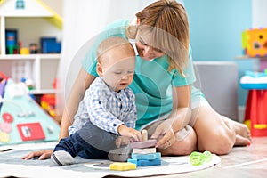 Happy mother and adorable baby boy playing on floor mat in sunny nursery room. Cute child with carer in creche
