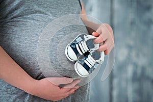 Happy mother in anticipation of the birth of her son. Pregnant woman holding baby booties sneakers on tummy background. Close-up