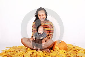 Happy mother and baby boy sitting on maple leaves by pumpkin