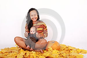 Happy mother and baby boy sitting on maple leaves by pumpkin