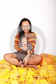 Happy mother and baby boy sitting on maple leaves by pumpkin