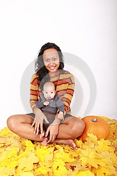 Happy mother and baby boy sitting on maple leaves by pumpkin