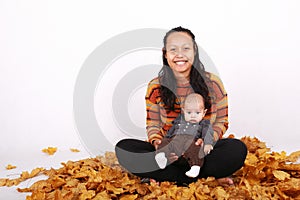 Happy mother with baby boy sitting on maple leaves by pumpkin