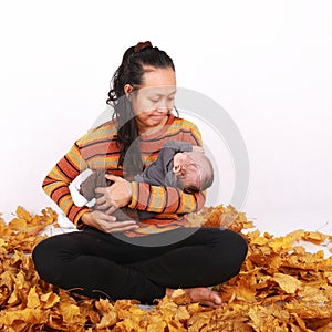 Happy mother and baby boy sitting on maple leaves by pumpkin