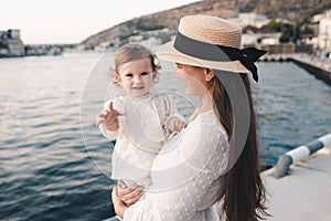 Happy mother and baby daughter having fun wear white stylish dress and straw hat over sea shore outdoors.