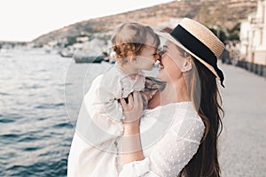 Happy mother and baby daughter having fun wear white stylish dress and straw hat over sea shore outdoors.
