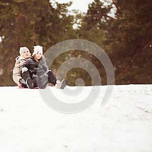 Happy mother with baby girl sitting on sledge and sledding down on snow from hill. Enjoying white winter day at park