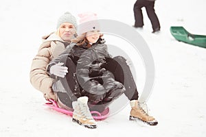 Happy mother with baby girl sitting on sledge and sledding down on snow from hill. Enjoying white winter day at park