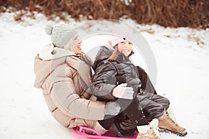 Happy mother with baby girl sitting on sledge and sledding down on snow from hill. Enjoying white winter day at park