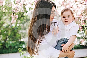 A happy mother with a baby in her arms in white clothes looks into his eyes against background of cherry blossom