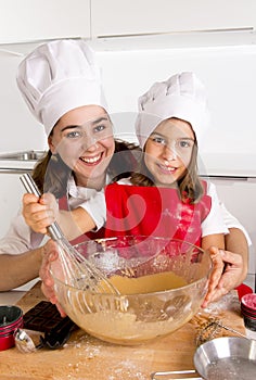 Happy mother baking with little daughter in apron and cook hat preparing dough at kitchen