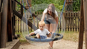 Happy mother and boy swinging in the round rope nest swing on the playground. Kids playing outdoors, children having fun, summer