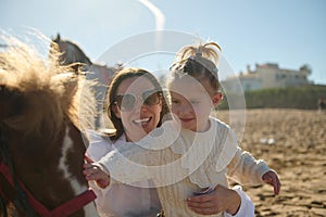 Happy mother carrying her daughter while stroking a little pony outdoor