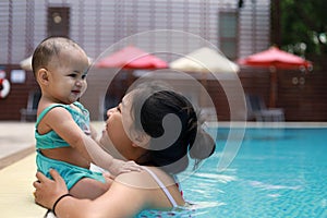 Happy mother and cheerful little baby girl enjoy playing together at outdoor swimming pool in long vacation. Family activity.