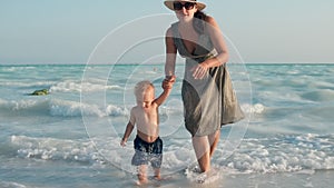 Happy mother and child going from water at seashore. Cute kid sitting in water.