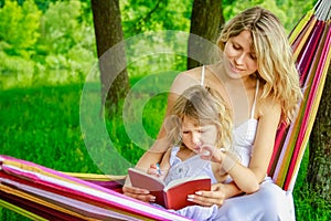 Happy mother with a child reading a book on a hammock in the nature park