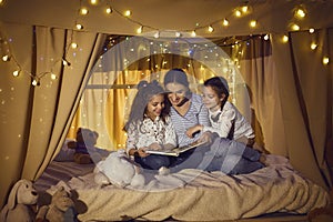 Happy mother and children sitting in playroom tent and reading book of fairy tales