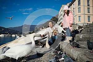 Happy mother and daughter feeding swans and ducks at the lake of Como in Italy, on a sunny cold winter day. Beautiful