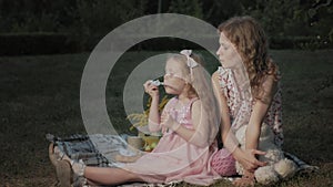 A happy mother and daughter inflate soap bubbles. Family in a city park on a picnic on a warm evening at sunset.