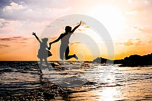 Happy mother and daughter jumping together along the sea water - Silhouette of loving family having fun on the beach