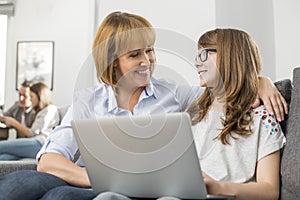 Happy mother and daughter with laptop while family sitting in background at home