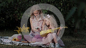 A happy mother and daughter are playing on the ukulele. Family in a city park on a picnic on a warm evening at sunset.