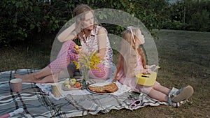 A happy mother and daughter are playing on the ukulele. Family in a city park on a picnic on a warm evening at sunset.