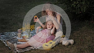 A happy mother and daughter are playing on the ukulele. Family in a city park on a picnic on a warm evening at sunset.