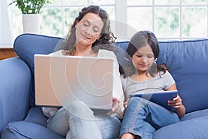 Happy mother and daughter sitting on the couch while using laptop and tablet