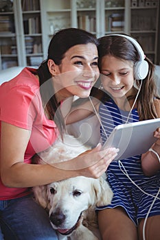 Happy mother and daughter sitting with pet dog and listening to music on headphones