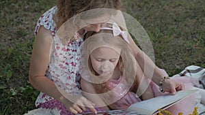 A happy mother and daughter view photos in an album read a book. Family in a city park on a picnic on a warm evening at