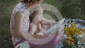 A happy mother and daughter view photos in an album read a book. Family in a city park on a picnic on a warm evening at