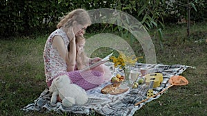 A happy mother and daughter view photos in an album read a book. Family in a city park on a picnic on a warm evening at