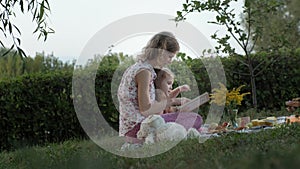 A happy mother and daughter view photos in an album read a book. Family in a city park on a picnic on a warm evening at