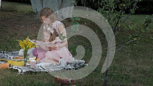 A happy mother and daughter view photos in an album read a book. Family in a city park on a picnic on a warm evening at