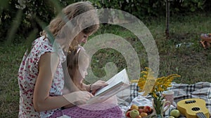 A happy mother and daughter view photos in an album read a book. Family in a city park on a picnic on a warm evening at