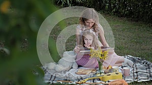 A happy mother and daughter view photos in an album read a book. Family in a city park on a picnic on a warm evening at