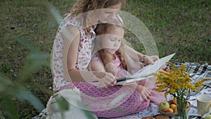 A happy mother and daughter view photos in an album read a book. Family in a city park on a picnic on a warm evening at